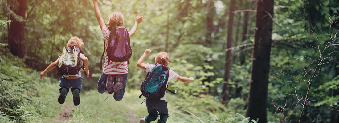 3 children wearing backpacks jumping for joy on a forest trail